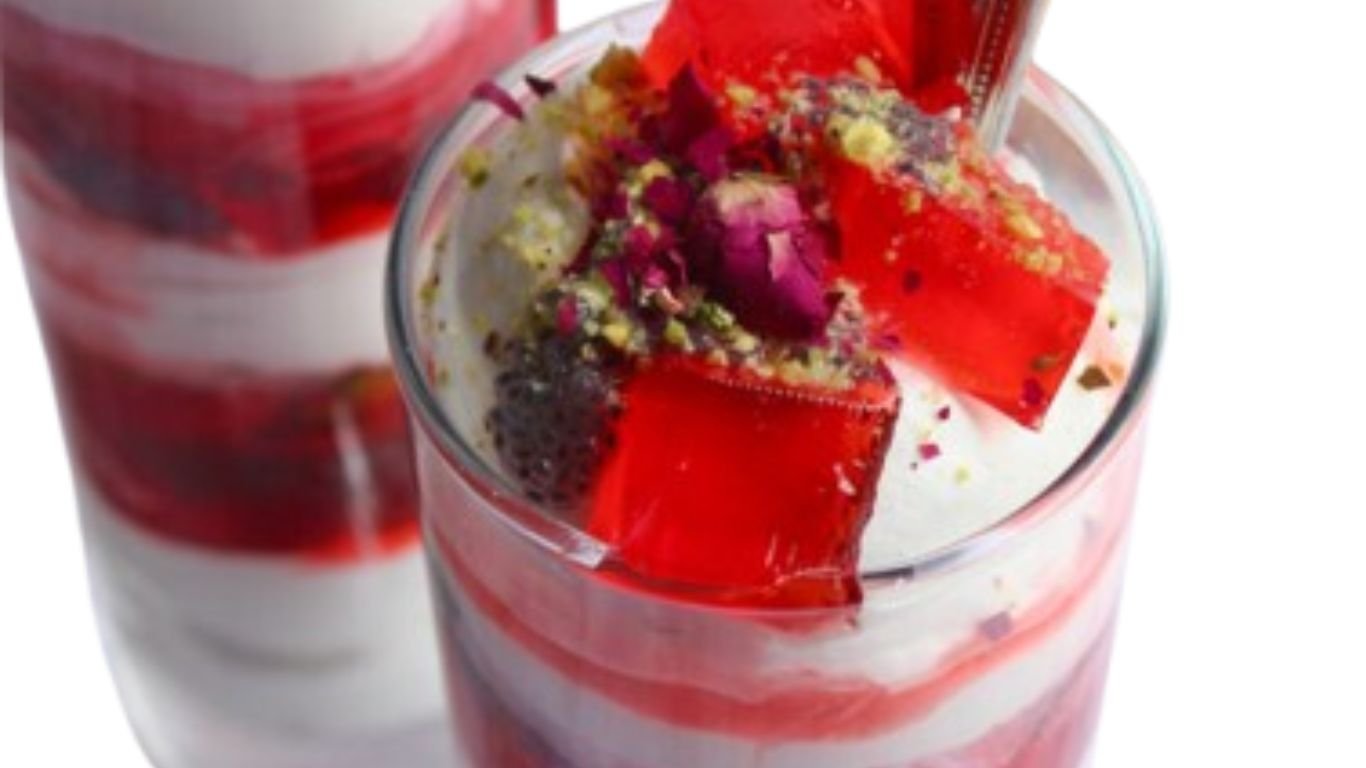 A delicious falooda dessert served in a tall glass, layered with rose syrup, milk, falooda sev, and sabja seeds, topped with red jelly cubes, crushed nuts, and dried rose petals